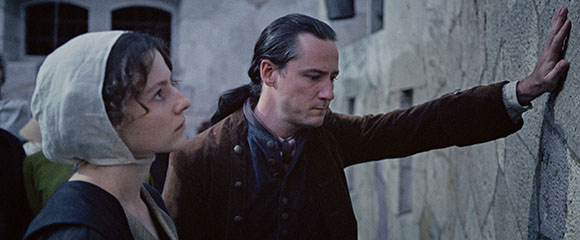 A man in a brown coat reaches toward a stone wall while a woman in a white headpiece looks on.