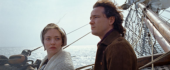 A man and a woman in period clothing stand on a wooden sailing vessel, looking toward the horizon.