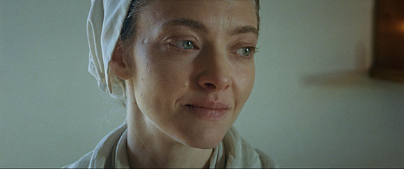 A woman wearing a white bonnet looks off-camera with a subtle, emotional expression.