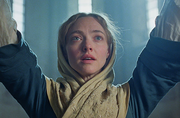 A woman in a headscarf looks upward with her arms raised against a hazy, bright background.