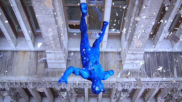 A person in a bright blue bodysuit falls headfirst through a shattering glass window of a building.