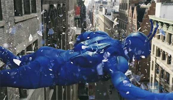 A person in a bright blue bodysuit falls through the air between city buildings amidst flying white debris.