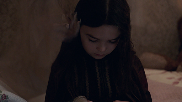 A young girl with long dark hair looks downward in a dark, moody setting.