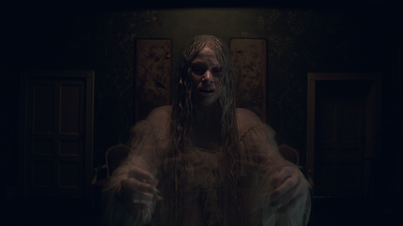 A pale woman with long, messy hair and a white gown stares intensely in a dimly lit room.