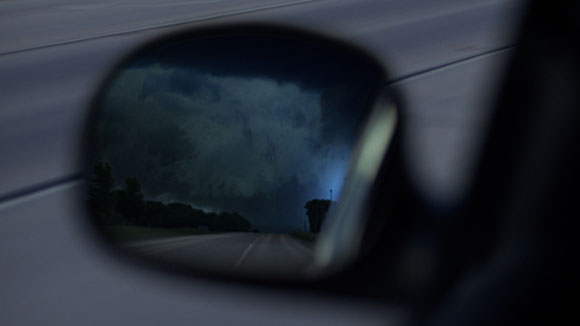 A car side mirror reflects a dark, cloudy sky over a road at dusk.