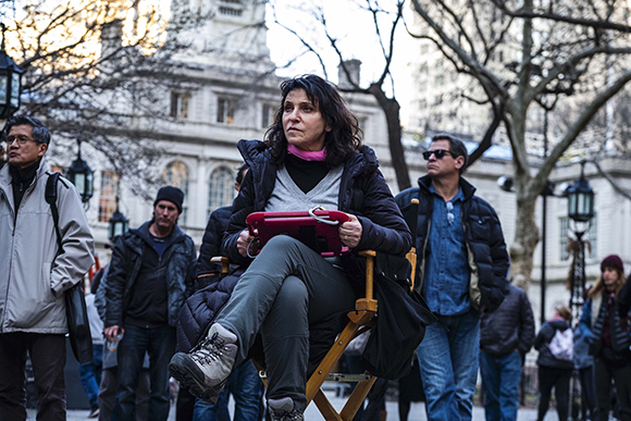 A woman in a dark jacket sits in a wooden chair holding a red tablet in a city setting.