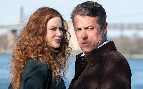 A woman with long wavy red hair and a man in a dark brown coat stand against a blurred river background.