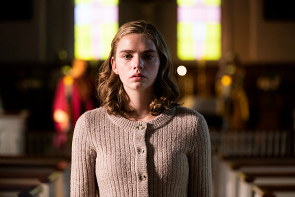 A young woman with wavy brown hair wearing a beige cardigan stands in a dimly lit church.