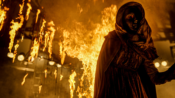 A person in a dark, hooded cloak stands in front of large, bright orange flames.