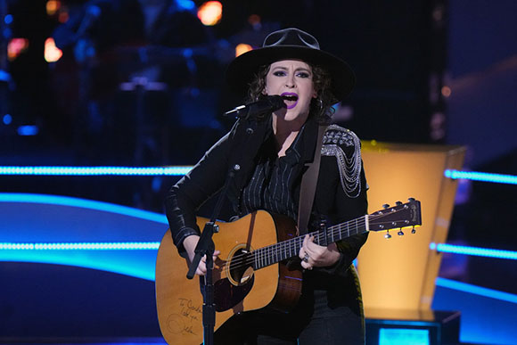 A woman in a black hat and jacket sings into a microphone while playing an acoustic guitar on a lit stage.