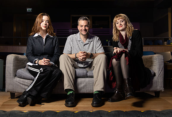 A man and two women sit together on a grey couch in a dimly lit room.