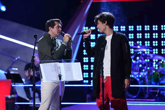 Two young men perform on a stage with microphones, music stands, and a blue light grid background.