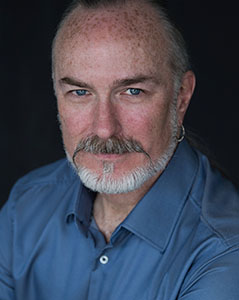 A man with light skin, blue eyes, a grey goatee, and a silver hoop earring wearing a blue button-down shirt.