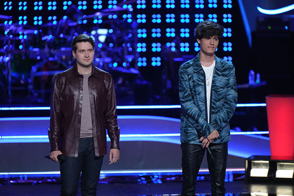 Two young men standing on a stage with blue lights and a dark background.