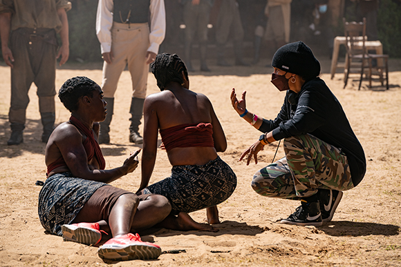 Two women sit on the dirt ground while a third person in a black beanie crouches and gestures toward them.