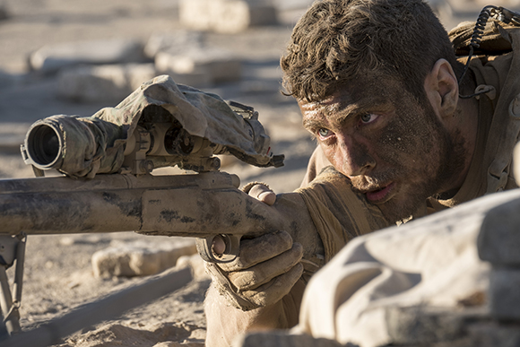 A dirt-covered soldier lies prone, looking through the scope of a long-range rifle in a desert environment.