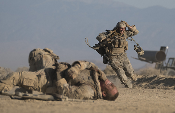 A soldier runs through a desert while another lies prone on the ground in the foreground.