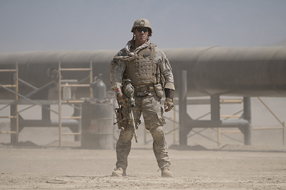 A soldier in camouflage gear and tactical vest stands in a dusty desert landscape near large industrial pipes.