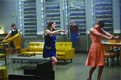 Three women in vintage dresses move around a room with a yellow sofa and large windows.