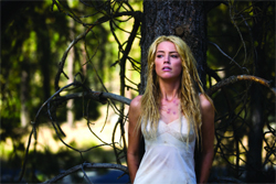 A blonde woman in a white tank top leans against a tree trunk in a sunlit forest.