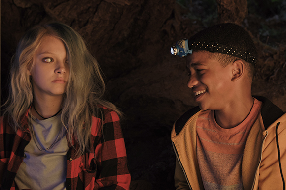 A young girl with blonde hair and a boy wearing a headlamp look at each other in a dark setting.