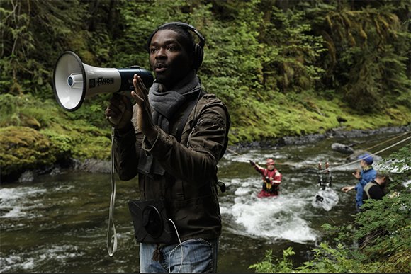 A man in a brown jacket and headphones holds a megaphone near a flowing river in a forest.