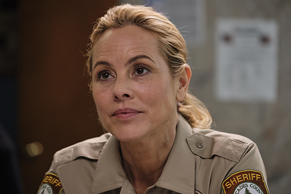 A blonde woman in a tan sheriff's uniform looks upward with a thoughtful expression.