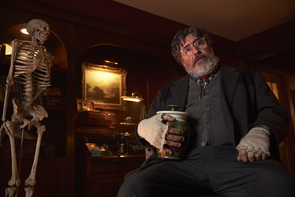 A bearded man in a suit sits in a dark room holding a jar next to a human skeleton.