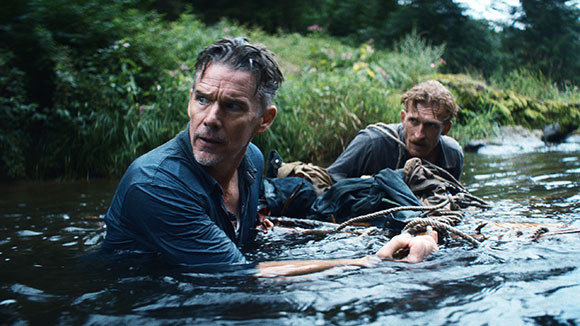 Two men in dark clothing wade through a river while carrying heavy gear and ropes.