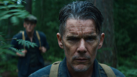 Close-up of a man with short dark hair and a stern expression in a dense forest.