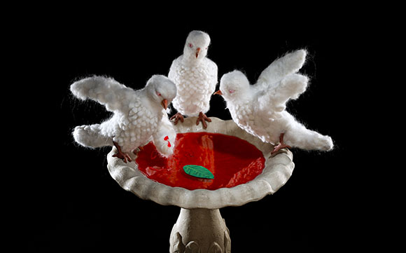 Three small white felted birds perched on the edge of a stone birdbath filled with red liquid.
