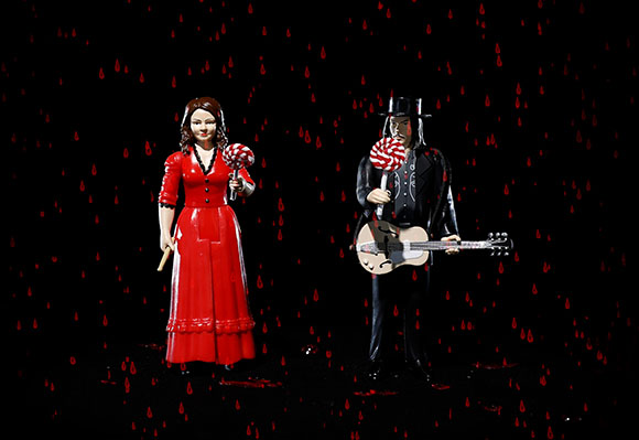 A female figurine in a red dress and a male figurine in a black suit with a guitar stand against red droplets.