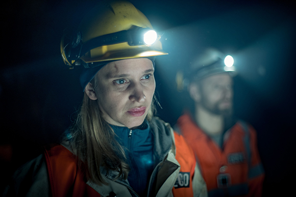 A woman in a yellow hard hat with a headlamp looks forward in a dark setting, with a man behind her.