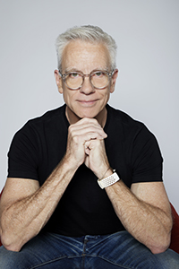 A man with white hair and glasses wearing a black t-shirt, sitting with his hands clasped under his chin.