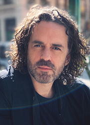 Close-up portrait of a man with dark, curly hair and a salt-and-pepper beard looking at the camera.