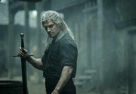 A man with long white hair and a dark shirt holds a large sword in a misty, dark setting.
