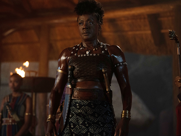 A woman with short dark hair wearing patterned armor and gold bracelets stands in a dark, torch-lit room.