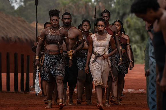 A group of people in traditional patterned clothing and armor walking through a village setting.