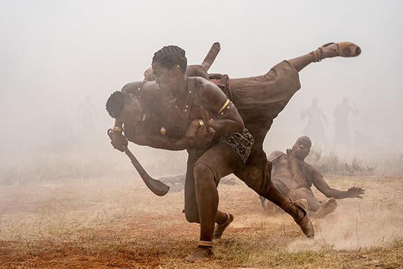 Two men in traditional attire struggle intensely in a dusty, foggy field.