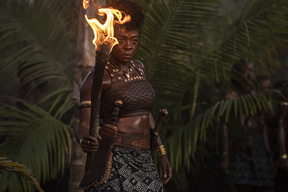 A woman in traditional warrior attire holds a flaming torch in a lush, tropical jungle.