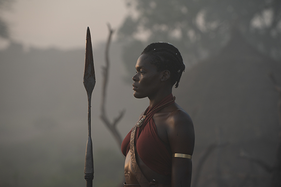 Profile view of a woman in traditional red attire holding a long spear against a misty landscape.