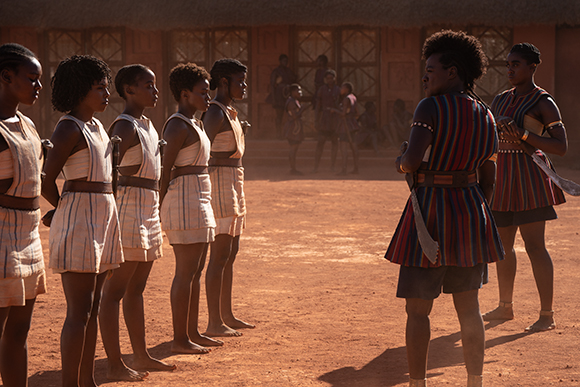 A line of women in light-colored tunics face a woman in a colorful striped tunic on a dirt ground.