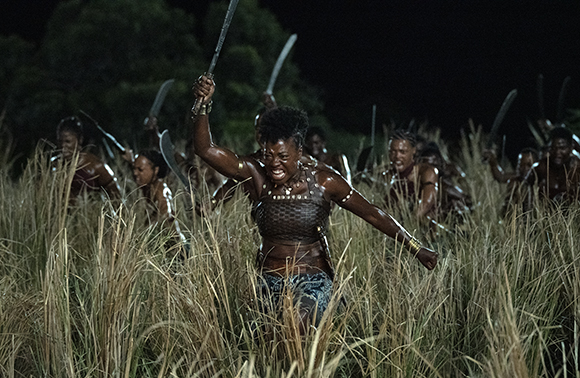 A woman in leather armor charges through tall grass at night, brandishing a sword with a group of warriors.