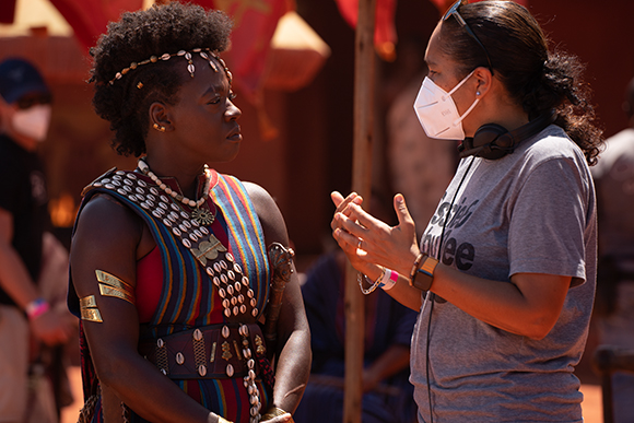 A woman in colorful patterned clothing talks to a woman wearing a white face mask and headphones.