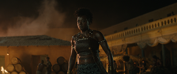 A dark-skinned woman in patterned warrior clothing stands in a dimly lit village at night.