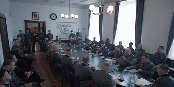 Military officers in uniform sit around a long dark table in a formal meeting room.