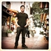 A man in a black t-shirt and dark pants stands in a narrow, sunlit street alley.