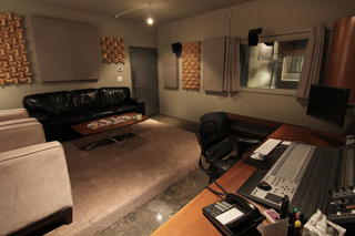 A recording studio control room featuring a large mixing console, a black leather sofa, and acoustic wall panels.