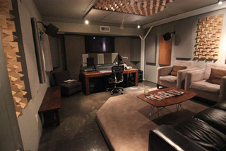 A music studio featuring a desk with monitors, acoustic wall panels, and a lounge area with armchairs.