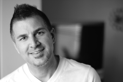 A smiling man with short dark hair wearing a white v-neck t-shirt.